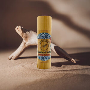 Baume à lèvres bio en tube sur sable, avec étiquette bois et motif bleu, boîte indication karité et calendula.