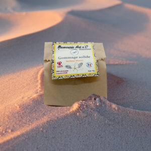 gommage solide Monoï posé sur un petit sac en papier, posé dans du sable avec lumière chaude._