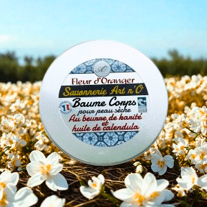 Baume corps Fleur d'oranger de 150 ml dans pot blanc aluminium au milieu de fleurs blanches en champ, étiquette bleue et dorée visible.